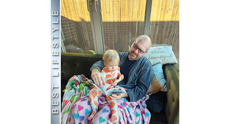 BEST LIFESTYLE: A father reading to his child, both covered with brightly colored throws, sitting on a sofa in a tan sunroom