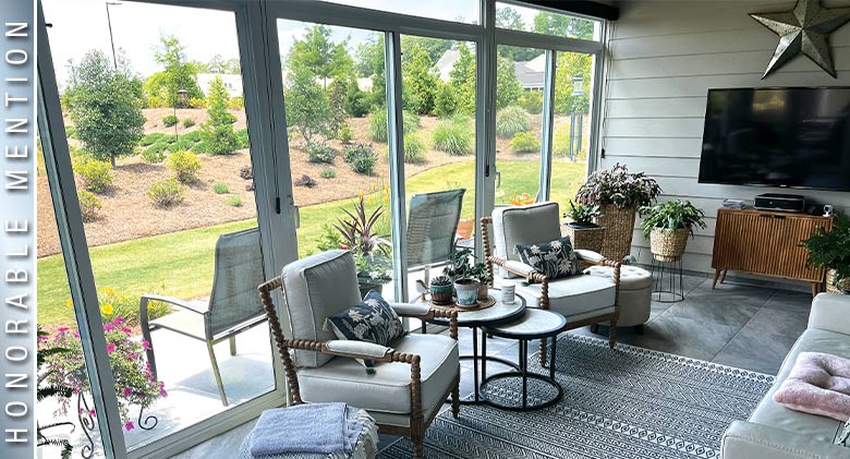 HONORABLE MENTION: Two chairs with light cushions, floral pillows, stacking tables and TV on wall of patio enclosure.