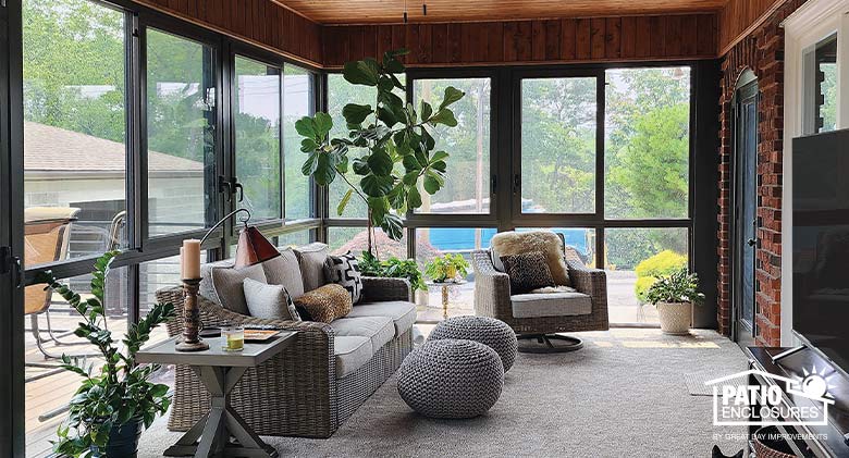 The interior of a brown sunroom with comfortable wicker seating with beige cushions, plants, wood ceiling, and TV on right.