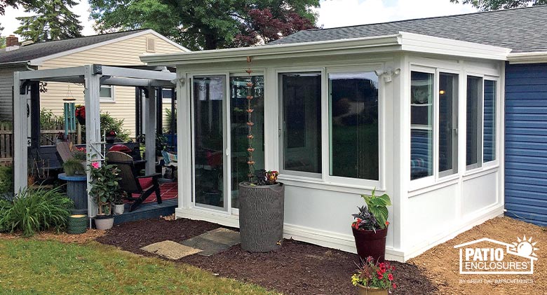 Exterior of white sunroom with shed roof and solid white knee wall, pergola, potted plants and rain barrel around exterior.