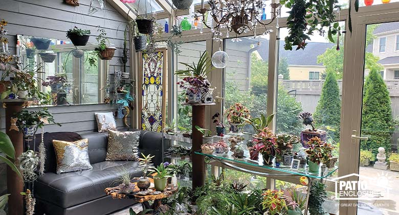 Interior of a solarium with plants hanging, on tables and on the floor. Mirror above gray leather sofa with
metallic pillows.