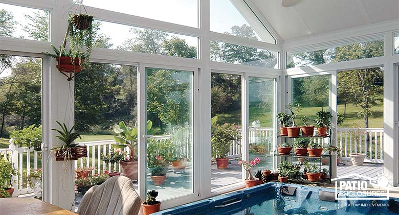 A white sunroom with soaring gable wall, a hot tub, and potted plants on a stand and hanging. More plants
outside on deck