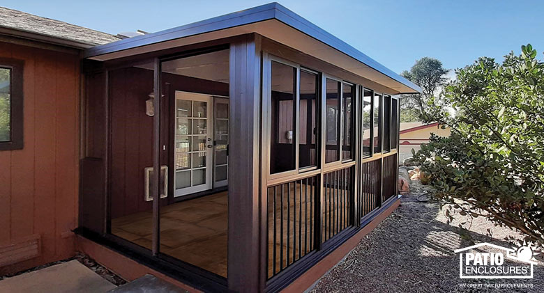 A brown screen room with picket railing, flat roof, and sliding screen doors on the end. Gravel and bushes in front.