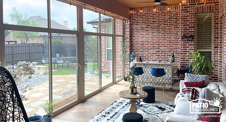 The interior of a glass sunroom with a small bar at the far end and a sofa and throw pillows on the right. The exterior brick wall of the home is also visible.