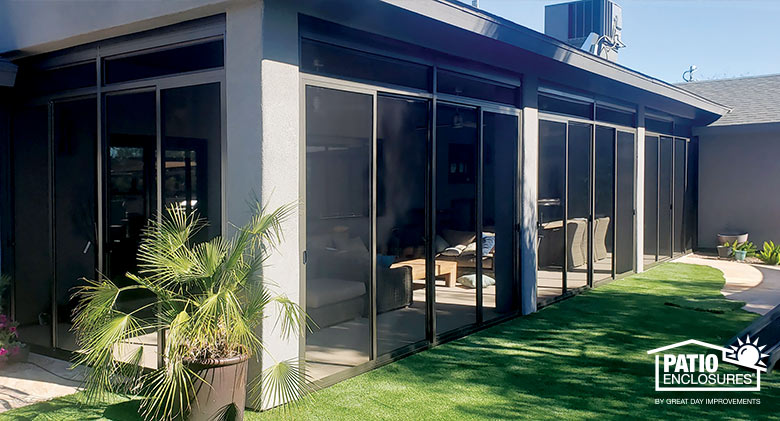 The interior of a glass sunroom with a small bar at the far end and a sofa and throw pillows on the right. The exterior brick wall of the home is also visible.