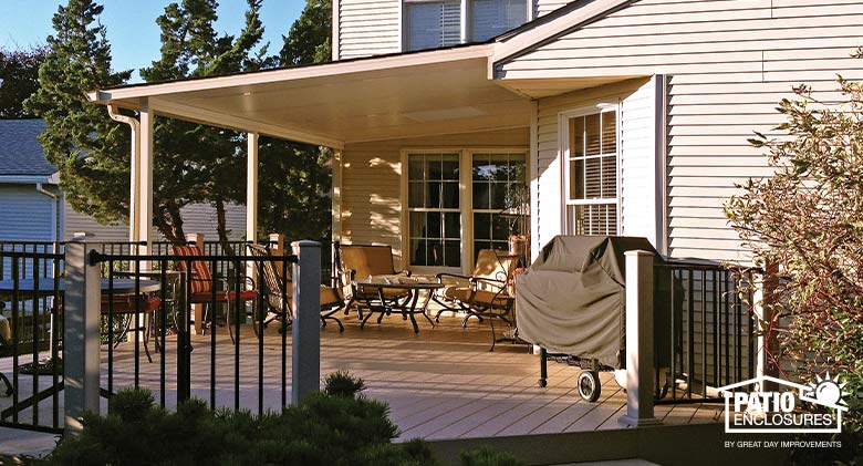 Paver patio covered with a tan roof, black railing around, tan and black chairs and grill.