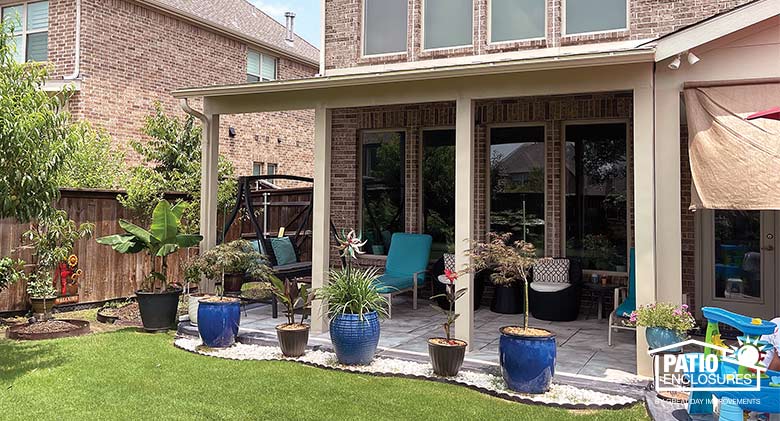 A patio on the end of a brick home with a tan cover, plants in blue and black pots in front of patio sitting on
white stones