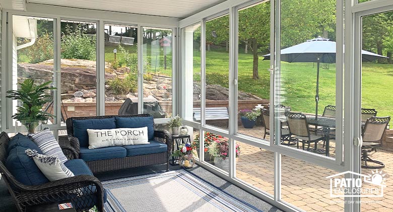 Sunroom patio enclosure with brown wicker furniture, blue cushions & pillow that says, “The Porch.” Table & chairs outside.