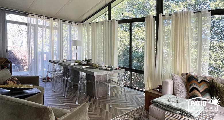 Sunroom interior with gable ceiling, dining table, conversation area with comfortable furniture, and sheer curtains.