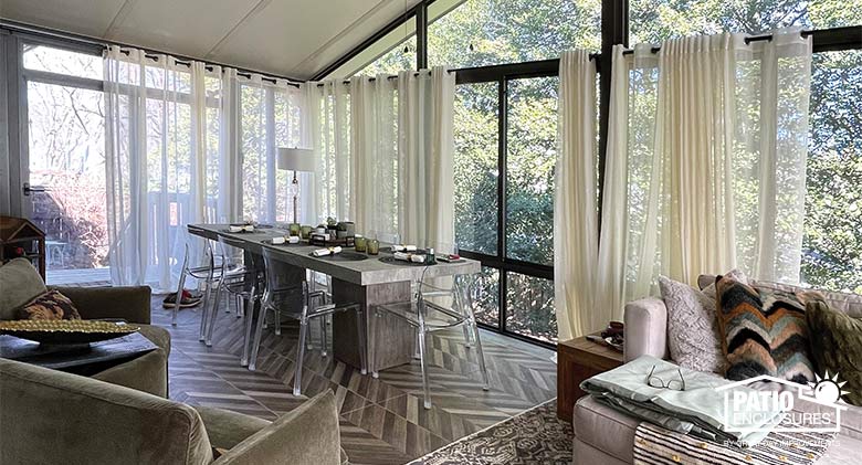  A large sunroom with gable roof featuring dining and seating areas and wispy, white sheer curtains partially closed