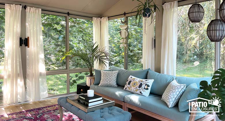 Sheer white curtains are opened to reveal the view from a tan sunroom with light blue upholstered furniture and plants.