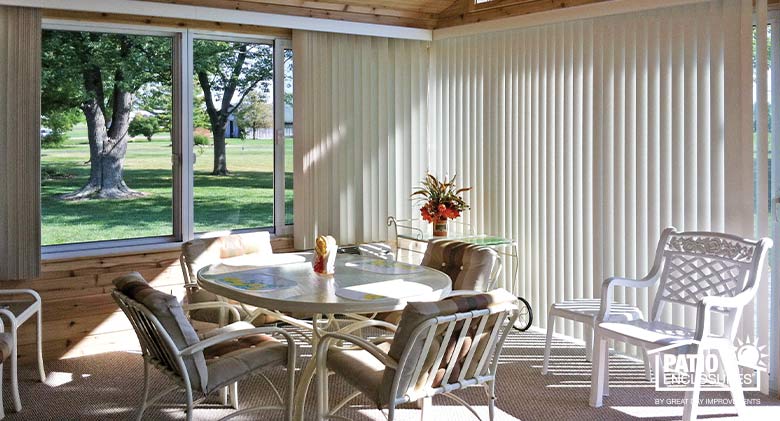 White vertical blinds in a sunroom with solid, paneled knee wall and furnished with tables and chairs.