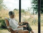 Woman with silver hair seated in a brown chair gazing out at the gardens through the glass of a sunroom.