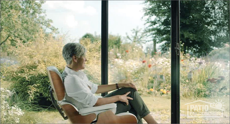 Woman with silver hair seated in a brown chair gazing out at the gardens through the glass of a sunroom.