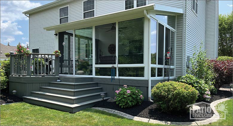 A white glass sunroom with a single-slope roof attached to a raised gray wood deck with neat landscaping around it.