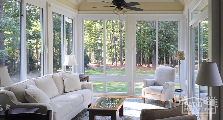 The interior of a glass sunroom with white frame furnished with a comfortable sofa, armchair, coffee table, and lamp