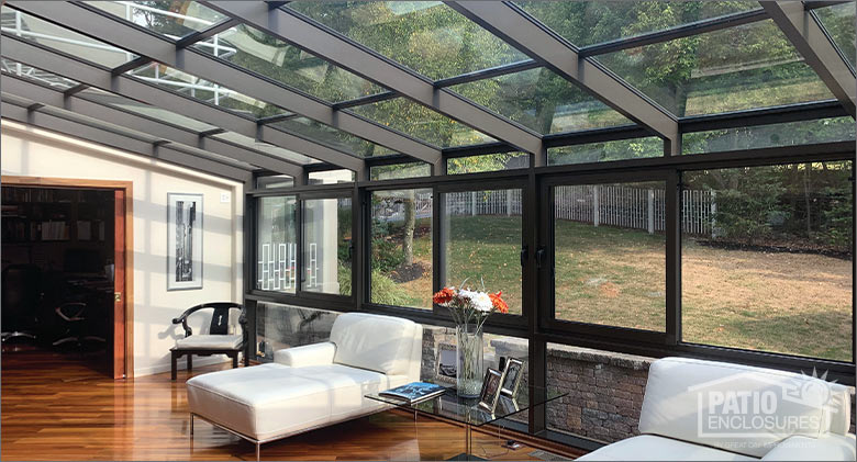 Two white upholstered chaises, an armchair, and a small table inside a brown solarium with glass roof and walls.