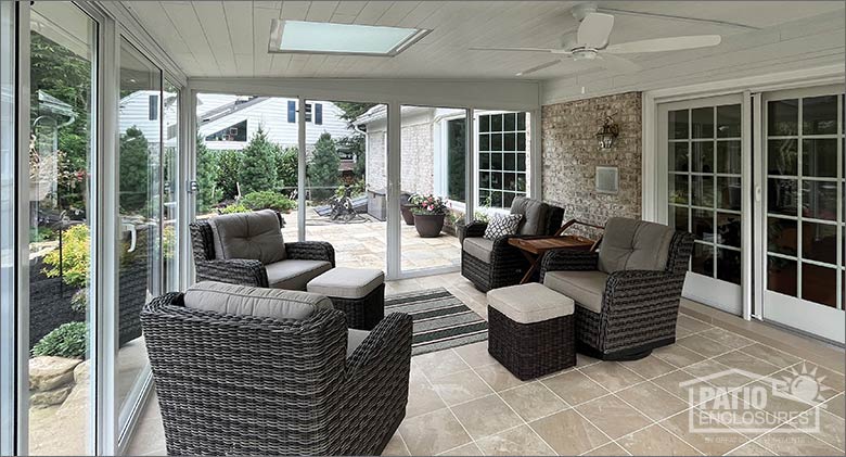 The interior of a sunroom with floor-to-ceiling windows, skylight, ceiling fan, and brown cushioned wicker furniture.