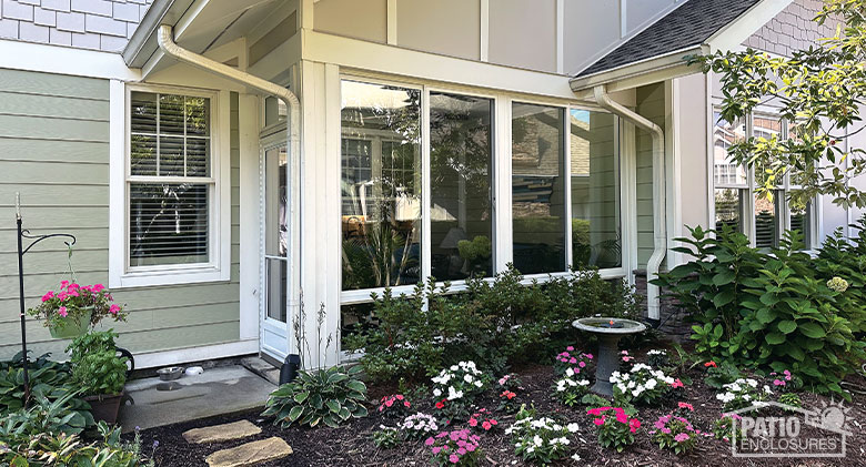 Sunroom Cost Photo Gallery A white two-walled patio enclosure with a storm door on the left side and white and pink flowers in front.