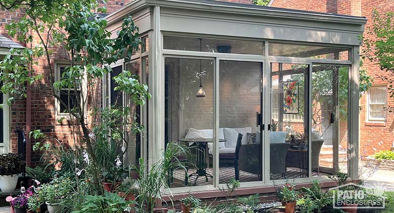 Sunroom Cost Photo Gallery A tan glass sunroom patio enclosure surrounded by potted plants and small trees. Inside, a comfortable seating area.