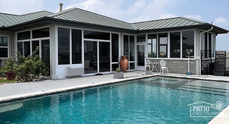 The exterior of an L-shaped patio enclosure with white frame and metal roof situated behind a large in-ground pool.