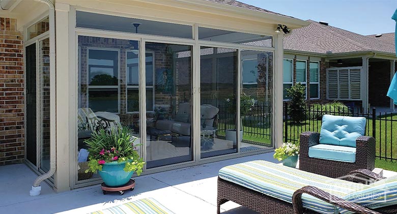 A sunny patio partially enclosed by a tan sunroom. Furniture with bright blue and green cushions, accented by potted plants.