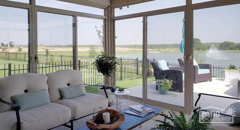 The shady interior of a glass patio enclosure with comfortable seating and view of a lake with fountain.