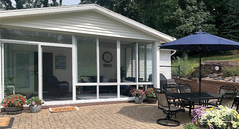 Exterior of a white patio enclosure with gable roof on a brick patio with table and chairs, blue umbrella, and potted flowers