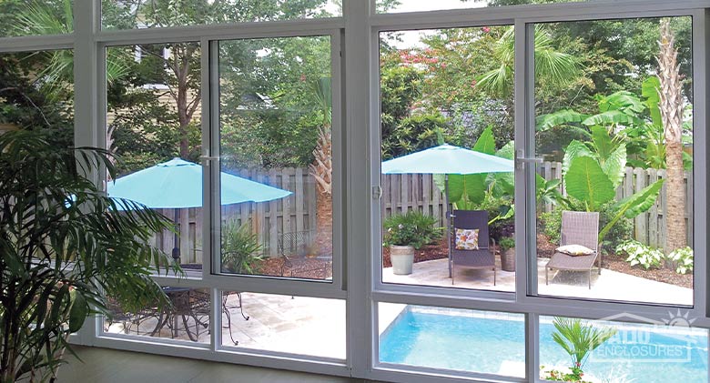  Interior of a glass sunroom overlooking a pool and patio with chaise lounges, a table, blue umbrellas, and lush plantings.