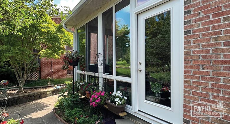 A white corner patio enclosure sunroom with potted flowers and a flower bed in front and a small tree on the side.