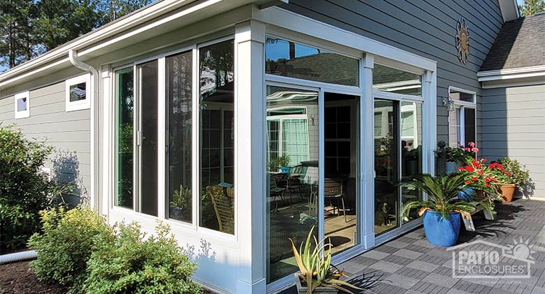 Corner patio enclosed in glass with many colorful potted plants outside on the gray patio and bushes on the side.