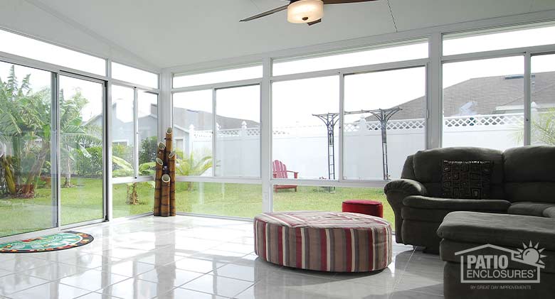 An overstuffed sofa and large ottoman furnish a white sunroom with white tile floor; stalks of bamboo decorate the corner.