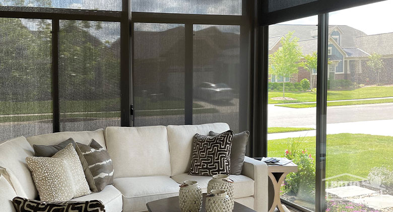 The interior of a brown sunroom with translucent shades pulled down on one wall and a white sofa with accent pillows.