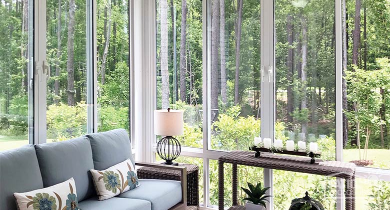 A light blue sofa with accent pillows and wicker side tables inside a white glass sunroom with glass knee wall.