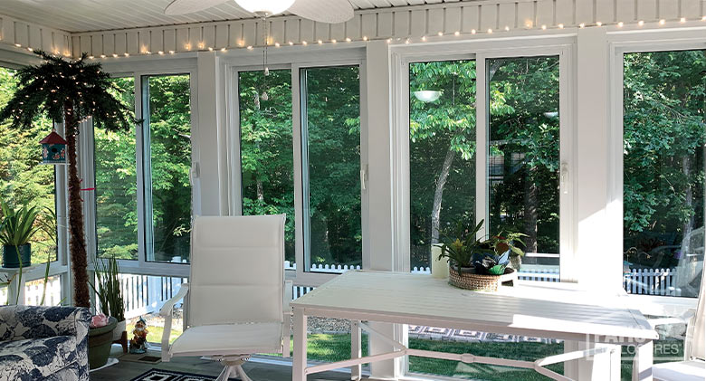 Mini white lights and lighted palm tree decorate a glass sunroom with table, chairs, and a glass knee wall.