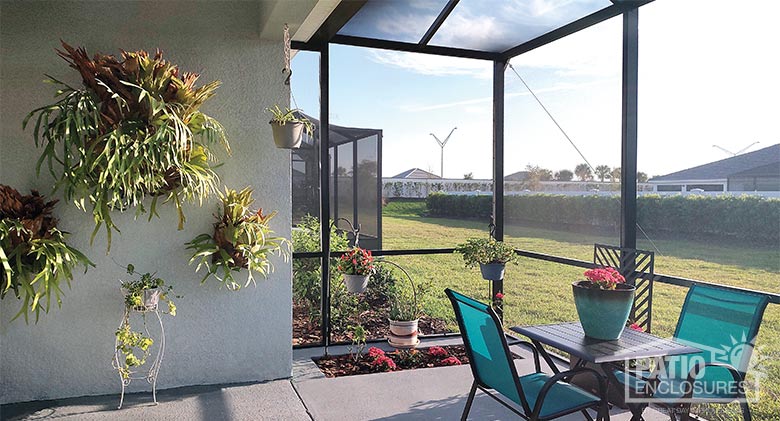 A table for two with blue chairs on a patio enclosed by screened walls and roof attached to a stucco home.