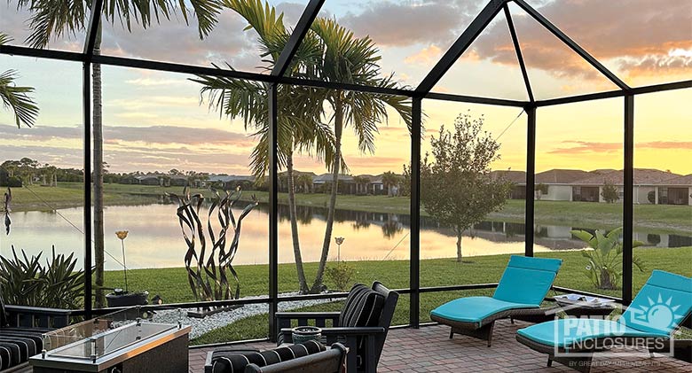 View of a sunset and small lake from inside a screen room with bright blue chaises, fire table, and other brown chairs. 