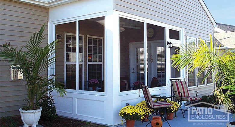 Striped cushions on two chairs & potted yellow mums sit on the patio in front of a white screened room with solid knee wall.