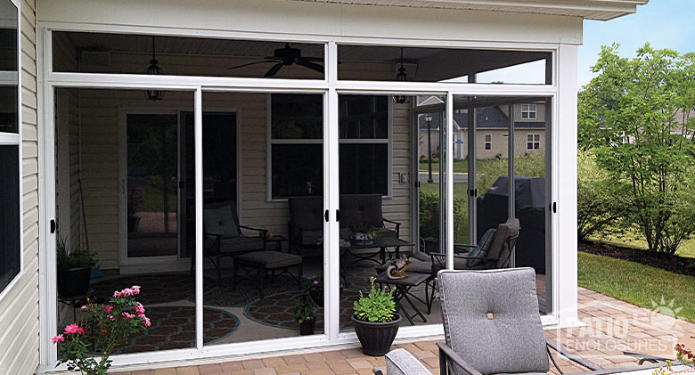 A corner patio enclosed with screens and a view inside of a seating area. Outside a grill, bushes, and potted plants.