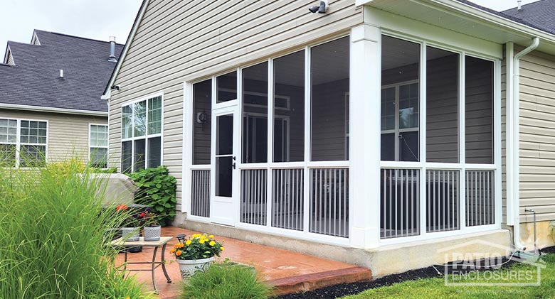 A white screen room with screen door and picket railing with a brick patio, potted flowers, and grasses in front of the room.