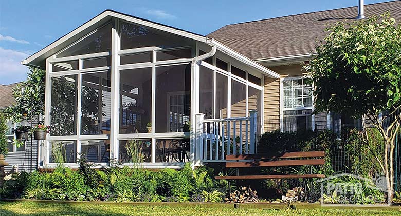 A large white screen room with gable roof surrounded by lush plantings and a park bench.