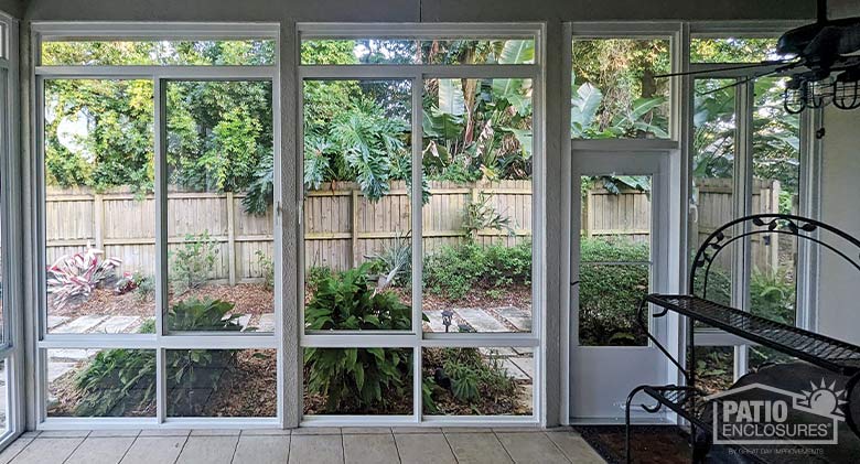 Looking out of a glass lanai enclosure onto gardens with stone paver paths and a wooden fence backdrop.