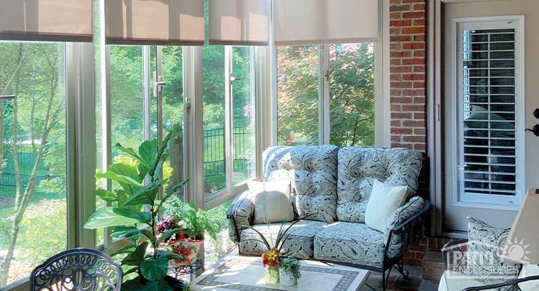 Sun streaming in through the glass of a porch or lanai enclosure with comfortable furniture and potted plants.