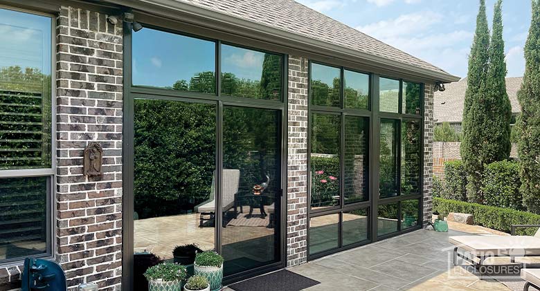 A lanai or porch on a brick home enclosed with glass walls with a brown frame, patio furniture and potted plants outside.